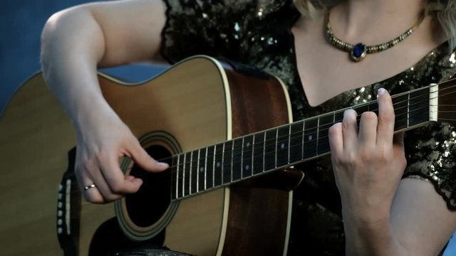 A girl with a necklace on her neck plays an acoustic guitar. The right hand touches the strings, the left hand holds the chords on the fingerboard. Smoke in the background. Day of music.