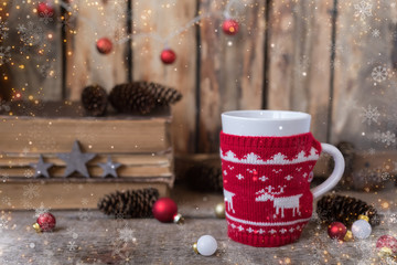 Knitted red cup with white reindeer, christmas lights with old books, cones and toys at background