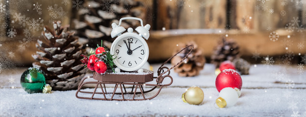 Christmas and New year alarm clock with snow on wooden sled with cones and toys, banner