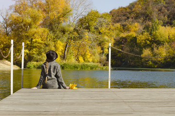 Girl in the hat sitting on the dock. Autumn day. Back view