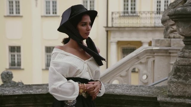 Pretty young model in extravagant boho style look seductively poses to camera outdoors, standing on an ancient balcony. Professional photoshoot, celebrity, top model. Female portrait