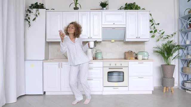 Young Excited Woman Dancing In Pajamas Holding Smartphone In Kitchen.