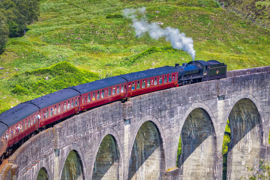 Close shot of Jacobite train travelling over the Glenfinnan Viaduct with a green hillside behind it and steam coming out of the locomotive
