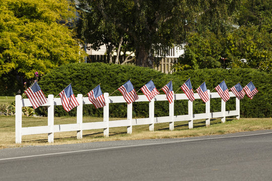 Patriotic display of American flags waving on white picket fence next to a road. Typical small town Americana Fourth of July Independence Day decorations.