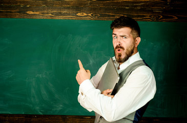 Education, high school, technology concept - teacher standing with laptop computer in front of chalkboard and shows finger on blackboard. Back to school concept. Surprised male teacher in classroom.
