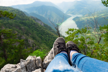 The view of female legs and feet in jeans and black leather tennies shoes on a rock with mountains...