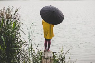 girl stay on small wooden pier under black umbrella in yellow raincoat back to camera in outdoor...