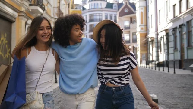 Group of three multiethnic teenage female friends walking in shopping area in city holding shopping bags. Beautiful mixed race girl smiling and hugging her asian and caucasian girlfriends