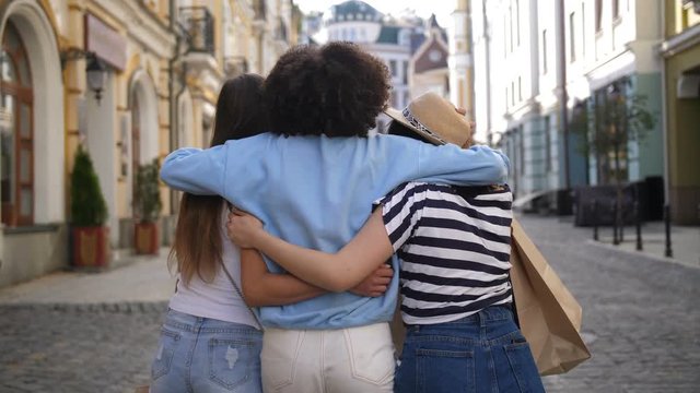 Rear view of multiracial girlfriends walking with shopping bags and embracing on street. Cheerful asian, mixed race and caucasian young female friends hugging and celebrating successful purchases