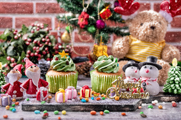 set of Christmas celebration gifts, cup cake, sugar santa , sugar, snowman, bear doll with background of Christmas tree in front of brick wall with falling snow