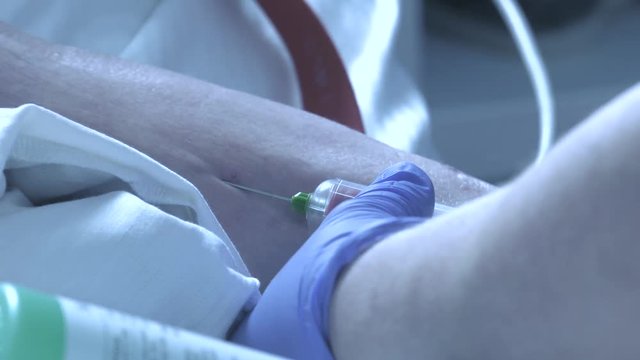 An elderly man lies on a bed in a hospital. The nurse takes blood for a test with a syringe and a needle. In blue gloves imposes a patch.