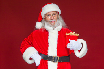 Christmas. Smiling, kind Santa Claus in white gloves holds a red bucket with popcorn with one hand, and puts the other hand on his belt. The concept of visiting the cinema, watching a movie with