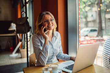 Cheerful young beautiful woman with glasses talking on mobile phone and using laptop with smile while sitting at her working place