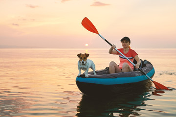 Red-haired young woman is rowing on an inflatable kayak by the sea with a dog Jack Russell Terrier on a background of pink sunrise in beautiful nature. Great disk of the rising sun. Sun rays. Sport - obrazy, fototapety, plakaty