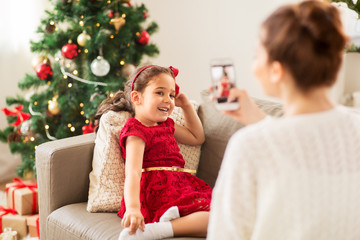 christmas, technology and family concept - mother photographing little daughter by smartphone at home