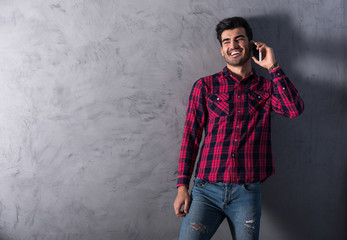 Fototapeta na wymiar A happy handsome young man in a red checkered shirt talking on his phone in front of a grey wall in a studio.