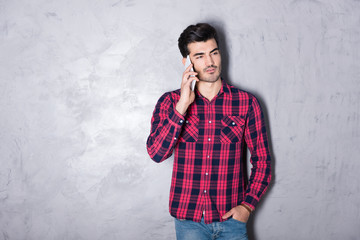 A serious handsome young man in a red checkered shirt talking on his phone in front of a grey wall in a studio.