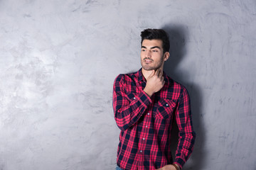 A thinking handsome young man in a red checkered shirt standing in front of a grey wall in a studio.