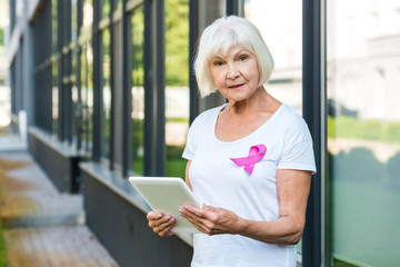 senior woman with pink ribbon on t-shirt using digital tablet and looking at camera, breast cancer...