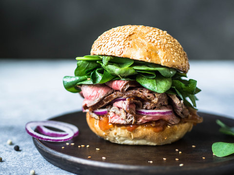 sandwich with steak slices, sauce of salsa, spinach and red onion. Burger from a bun with sesame and steak on a wooden plate on a gray background
