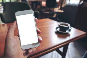 Mockup image of hand holding white mobile phone with blank desktop screen with coffee cup on wooden table in cafe