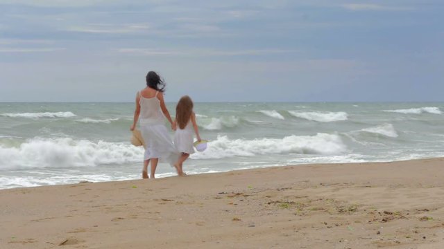 Mom and his beloved daughter are walking along the seashore, holding beach hats and chatting