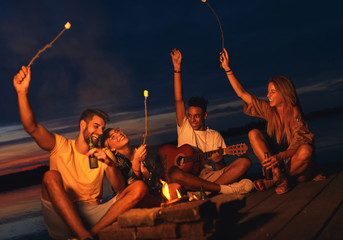 Group of young friends enjoying at the lake at night. They sitting around the fire singing and having fun.