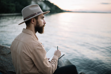 Portrait of young bearded man writes notes in a notebook, enjoying beautiful nature and sunset....