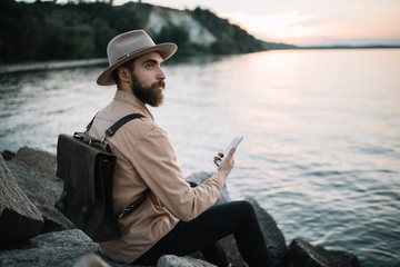 Portrait of young bearded man holding mobile phone, admire beautiful nature and sunset. Handsome traveller wearing hipster hat and leather backpack sitting on the river bank. Time to travel concept.