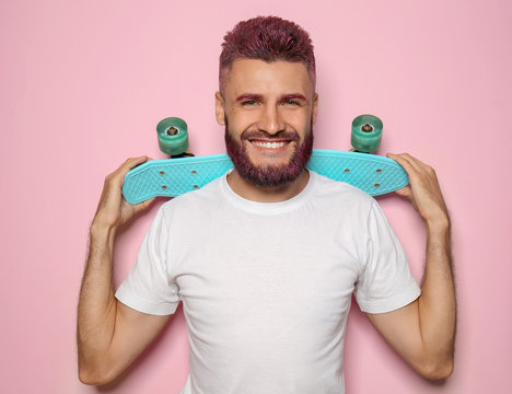 Portrait of handsome man with dyed hair and beard holding skateboard on color background