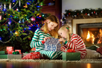 Fototapeta na wymiar Happy little sisters wearing Christmas pajamas playing by a fireplace in a cozy dark living room on Christmas eve.