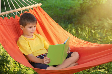 Cute little boy reading book while relaxing in hammock outdoors