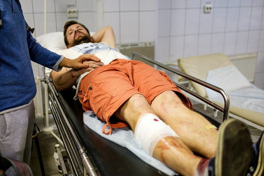 Young Man Lying In A Hospital Bed In The Building Of Ambulance Of A Doctor With An Injured Leg Bloodied And Temporary Bandaging After A Car Accident. Nearby Stands A Woman And Holding His Hand.