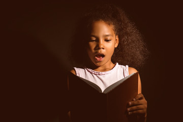 Surprised African-American girl reading book on dark background