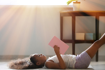 Little African-American girl reading book while lying on floor at home