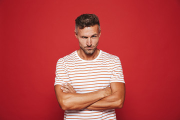 Irritated man 30s in striped t-shirt standing with arms crossed and angry look, isolated over red background