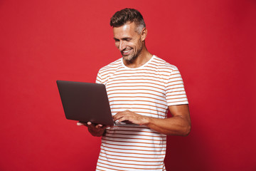Image of brunette man in striped t-shirt smiling and holding gray laptop, isolated over red...
