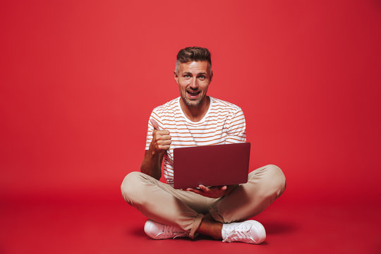 Image Of Adult Man 30s In Striped T-shirt Smiling And Using Gray Laptop, While Sitting On Floor With Legs Crossed Isolated Over Red Background