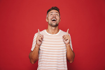 Happy man in striped t-shirt gesturing index fingers upward at copyspace, isolated over red...