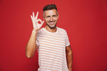 Brunette young man in striped t-shirt smiling and showing ok sign, isolated over red background