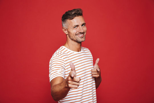 Caucasian man 30s in striped t-shirt gesturing index finger on camera meaning hey you, isolated over red background