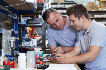 Engineer Helping Male Apprentice In Factory To Measure Component Using Micrometer