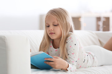 Cute little girl reading book on sofa at home