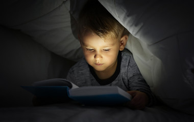 Cute little boy reading book in bed under blanket - Powered by Adobe