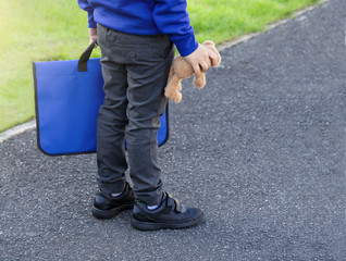 Cropped shot of schoolboy carrying school bag and teddy bear standing on street,View of legs with black leather school shoes worn by a boy student standing on cement path,