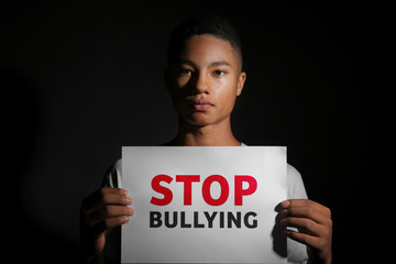African-American teenage boy holding sheet of paper with text STOP BULLYING on dark background