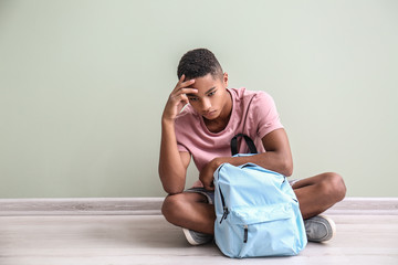 Sad African-American teenage boy sitting on floor indoors. Bullying at school