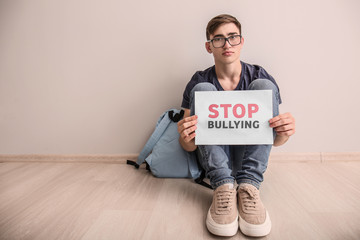 Teenage boy holding sheet of paper with text STOP BULLYING while sitting on floor indoors