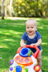 Cute little boy learning to walk with walker toy on green grass lawn at backyard. Baby laughing and having fun making first step at park on bright sunny day outdoors. Happy childhood concept
