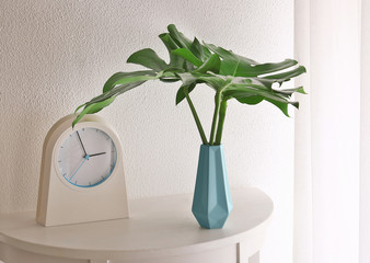 Tropical leaves in vase with clock on table near light wall indoors